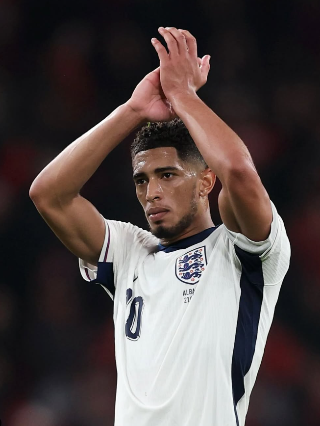 An image of Jude Bellingham celebrating after scoring a goal for Real Madrid, wearing the iconic white jersey and raising his arms to a cheering crowd at Santiago Bernabéu.